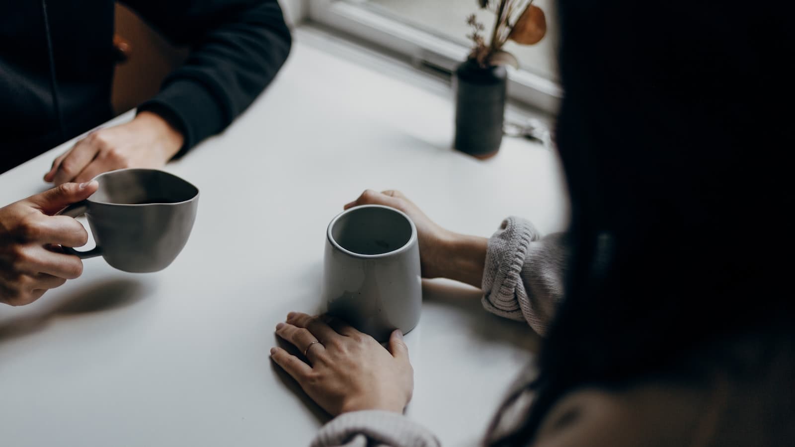 Two people at a bright white table, each holding a mug of coffee mid-conversation, with a small vase of dried botanicals by the window.