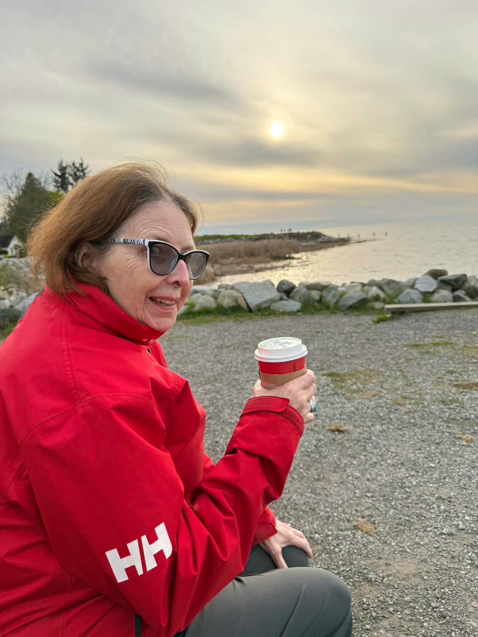 The senior in Samantha's life sitting at a beach at sunset, holding a coffee.